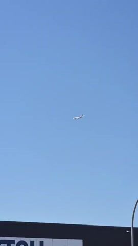A commercial airplane with white fuselage and red accents on its tail flies against a clear blue sky. The airplane is positioned in the upper center of the frame, moving from left to right. The bottom portion of the frame is occupied by the dark roof of a building.