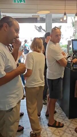 Here is a description of the video:

The video shows a bustling scene inside a cafe. A group of people are waiting in line near the cashier counter, while employees are busy preparing and serving orders behind the counter. One employee uses a point-of-sale system, while another prepares a drink using an espresso machine. The cafe's interior is visible, with artwork and decorations on the walls, and a peek into the kitchen area shows additional employees working with cooking equipment. Personal touches like children's drawings and photographs are also visible on a refrigerator.