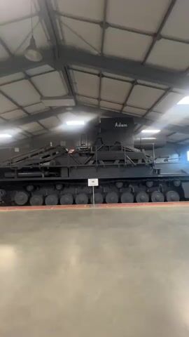 The video pans across a large, well-lit warehouse with a high, industrial ceiling. The floor is a smooth, gray concrete. A large, black, tracked vehicle, possibly a piece of construction or military equipment, is visible on the left side of the frame. It has the word "Adam" written on its upper section. Further into the warehouse, several other vehicles, appearing to be tanks or other military vehicles, are parked in rows. Some are dark green, and one has a white star emblem. The camera movement is smooth, suggesting a deliberate filming of the exhibit. The overall impression is of a museum or storage facility for heavy machinery.