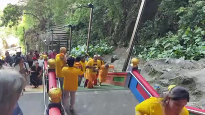 The video shows a group of people climbing a colorful staircase in a lush, outdoor setting. Many of the people are wearing yellow or orange clothing, and some are carrying small, decorative pots or containers. The staircase has a red, blue, and green railing with golden decorative elements. The surroundings are green, with vegetation covering the hillside and large rocks visible. Other people are also present, some of whom are also climbing the stairs. The group appears to be walking up towards a higher level, possibly a temple or other religious site, as they are chanting "Vel Vel." The video captures the movement of the group and the sounds of their chanting, highlighting the cultural and possibly religious nature of the scene.