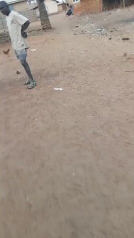 The video shows a man in a light-colored shirt and shorts walking on a dirt path. The camera follows him from a close perspective, then focuses on the ground. The background shows simple buildings and scattered debris. The environment appears to be a rural or less developed area.