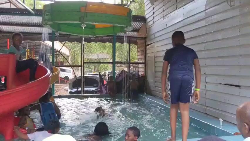 The video shows a group of children and adults at an indoor pool with a slide and overhead sprinkler system. A boy in a dark blue shirt and navy shorts stands at the edge of the pool, then performs a backwards flip into the water. Other people are in the pool, some swimming and playing near a red and yellow slide. One child is sitting on top of the slide. The pool is surrounded by a wooden fence, and a corrugated metal roof covers the area. Outside the enclosure, cars are parked.