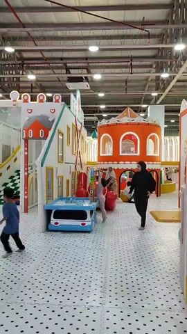 The video shows the interior of a children's indoor playground. The floor is covered with white foam tiles with black polka dots. In the foreground, a person wearing dark clothing walks from left to right. To the left of the frame, there is a colorful play structure designed to look like a house with a red roof and windows. In front of this structure, there is a blue and white play vehicle with a table on top, where two children are playing with red balls. To the right of the frame, there is a large orange structure with arched windows and a pointed roof, resembling a castle or tower. Other play areas and structures are visible in the background, as well as ceiling lights and exposed industrial ceiling elements. The overall environment appears bright and designed for children's entertainment.