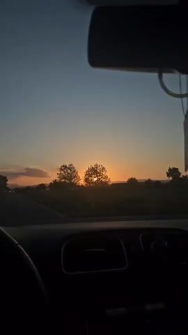 The video shows a view from inside a car driving towards a sunset. The sun, a bright orange orb, is partially obscured by trees in the distance. The sky above is a gradient of blue to orange, indicating twilight. A road stretches ahead, with occasional glimpses of buildings and distant hills. The car's dashboard is visible in the foreground, dimly lit, along with a portion of the steering wheel on the left side of the frame. The rearview mirror and a dangling object are also visible at the top of the frame. The car appears to be moving forward on a paved road. An electronic music track plays in the background.