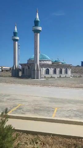 This video pans across a construction site or an industrial area under a clear blue sky. In the foreground, there is a paved area with yellow parking lines and a concrete curb, with dry soil and sparse vegetation. As the camera pans to the left, a mosque with a minaret and blue domes is visible. Further to the left, there is a tall tower-like structure, possibly a crane. The scene then shifts to show several long, rectangular buildings in the middle ground. As the camera continues to pan, large, domed industrial structures come into view, followed by a complex industrial facility with towers, conveyor belts, and silos in the background. The overall impression is one of development or ongoing industrial activity.