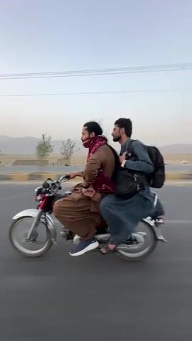 Two men are riding on a red motorcycle. The man in front is wearing a brown outfit and a red scarf. The man behind him is wearing a blue outfit and a black backpack. They are riding on a paved road with mountains in the background. The sky is clear and the sun is low in the sky, suggesting it is either sunrise or sunset.