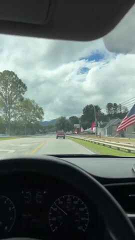The video shows a view from the driver's seat of a car looking forward through the windshield. The car is on a paved road with yellow dividing lines. A red pickup truck is in front, driving in the same direction. To the right of the road, there is a grassy area with a white guardrail, a few buildings, American flags, a large billboard, and some construction equipment. Trees and hills are visible in the background under a cloudy sky. The dashboard of the car is visible in the foreground.