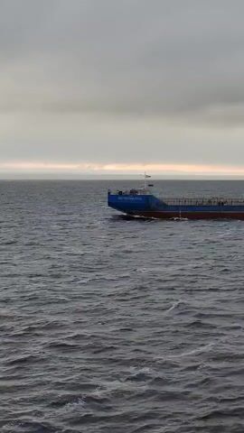 The video shows a blue and maroon ship sailing on a choppy sea under a cloudy sky. The ship, which has the name "MOZYRNA 3508" painted on its bow, is moving from right to left across the frame, leaving a white wake behind it. The sea is a dark gray, and the sky is a mix of gray and pale yellow clouds, hinting at a distant horizon. The camera seems relatively stable, providing a clear view of the ship as it crosses the water.