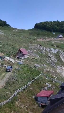 The video shows a rural mountainous landscape with several small wooden structures and a dirt path. A silver sedan is parked on the path. There are cows grazing on the grassy slopes. A few buildings are visible, some with red roofs. The sky is clear and blue.