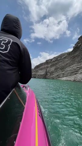 A video shows a person sitting on a hot pink and yellow boat, heading down a waterway with light green water. The person is wearing a black hooded jacket with a white number 83 on the back. High rock walls line each side of the waterway. The sky is blue with white clouds. Music is playing in the background.