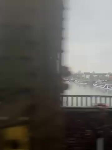 The video shows a view from a moving train crossing a bridge over a river on a cloudy day. Several tour boats are docked along the riverbank. Buildings are visible in the distance. A person walks along the pedestrian walkway of the bridge. The train is moving quickly.