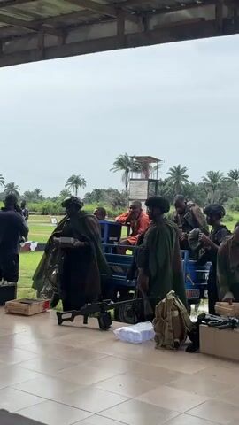 Several people are gathered outdoors under a sheltered area with a tiled floor.  Most wear military-style helmets, and several are wearing green ponchos. Rifles are propped against the side of a small utility vehicle that is loaded with several people. In the background are palm trees and a raised observation tower. Some individuals are handling rifles and opening boxes. One man in an orange shirt sits in the utility vehicle. Another man stands to the left, holding a notepad.