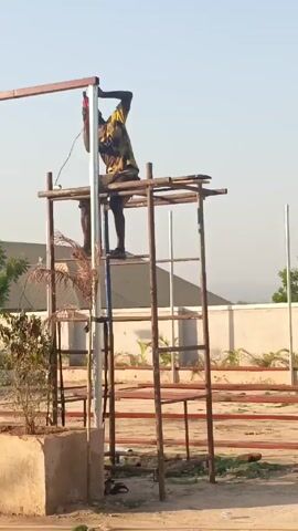 A man is using a power drill while standing on a rickety, makeshift scaffolding structure. He is wearing a dark t-shirt with a yellow pattern and dark shorts. The scaffolding is constructed from brown wooden beams and ladders, and it surrounds a white metal pole. There is a horizontal red metal beam above the man's head. The surrounding environment includes plants, a building with a dark roof, and a concrete wall. The sky is clear and light blue. The ground appears to be sandy.