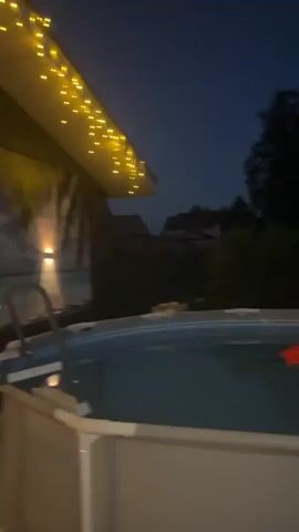 The video shows the exterior of a building at dusk. The building has a dark roof with yellow string lights hanging from the eaves. A backdrop featuring a beach and palm trees is attached to the wall of the building. There are two wall-mounted lights illuminating the backdrop. A security camera is mounted on the side of the building. A swimming pool with a ladder is visible in the foreground. Inside the building, a person is visible through a window, lounging on what appears to be a sofa or daybed. Someone speaks in the background. The overall ambiance is relaxed and inviting.
