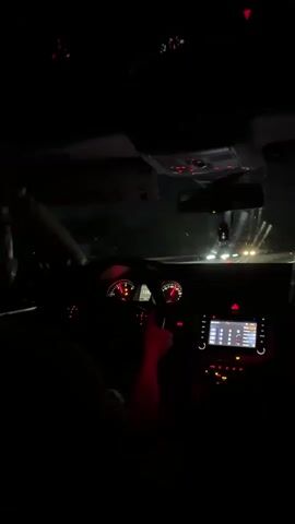 A person is driving a car at night. The interior is mostly dark, with illuminated dashboard gauges and the infotainment system screen. The driver's hands are visible on the steering wheel, and the car appears to be in motion on a road. The dashboard lights emit a red glow.