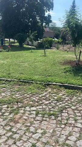 The video opens with a close-up shot of cobblestone paving, with small patches of grass growing between the stones. The camera then pans slowly to the right, revealing a grassy area with several small trees and some scattered flowers. In the background, there are several houses with red-tiled roofs, some appearing to be older and others newer. A few people can be seen walking in the distance, and a children's playground structure is visible. The sky is partly cloudy, with the sun shining through. The overall scene depicts a quiet, possibly rural or suburban, village setting on a sunny day.