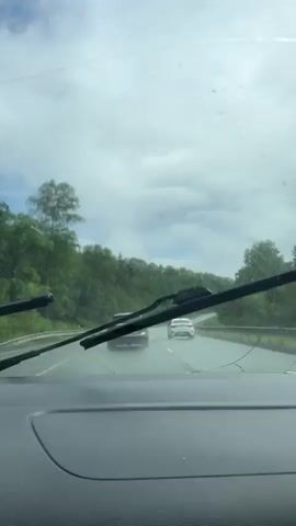The video shows a view from inside a car, looking out through the windshield. It is raining and the windshield wipers are in operation. Two cars are visible on a wet road ahead, driving in the same direction. The road is flanked by lush green trees and hills. The sky is overcast and cloudy. The video appears to be a continuous shot, likely taken while the car is in motion.