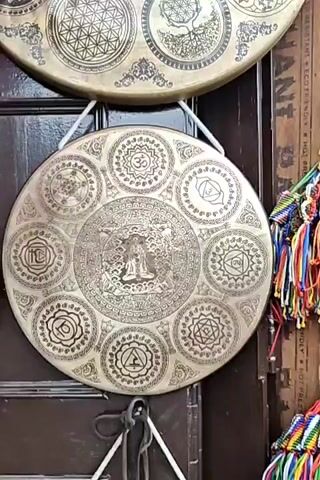 The video shows a close-up view of decorative gongs hanging against a dark wooden background. The gongs are made of a metallic material, likely brass, and are intricately engraved with various symbols and imagery. One gong prominently features a central image of a seated figure, possibly a deity or spiritual leader, surrounded by concentric circles of symbols including the Om symbol, chakras, and geometric patterns like the Flower of Life. Another gong displays a detailed engraving of a dragon's head in its center, also encircled by various symbols. A hand is seen briefly touching the surface of one of the gongs. The gongs are suspended by white ropes. In the background, a glimpse of colorful beaded items can be seen. The overall impression is of a display of ritualistic or decorative musical instruments.