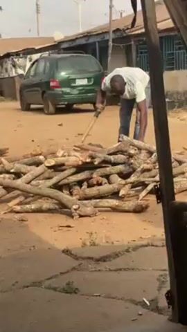 A man is splitting wood with an axe in an outdoor setting. He is wearing a white shirt and blue jeans. In front of him is a pile of branches and chopped wood scattered on the bare ground. Behind him, a green car is parked on the side of a dusty street, with buildings visible in the background. A wooden structure frames the lower right portion of the shot, partially obstructing the view.