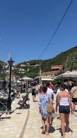 A vertical video shows a sun-drenched waterfront promenade. Several people are walking away from the camera, their backs to the viewer. To the left, several sailboats are docked in clear blue water. A lamppost stands on the promenade near the boats. In the background, buildings with red-tiled roofs are nestled on a lush, green hillside under a clear blue sky. The promenade is paved with light-colored stones.