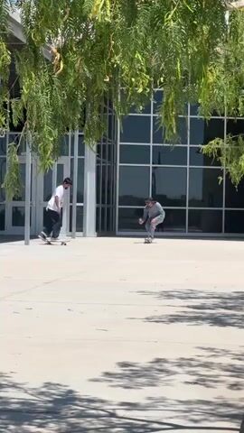Two males are skateboarding on a paved surface in front of a modern building with large windows. One male, wearing a white t-shirt and dark pants, is on the left side of the frame, looking down at his skateboard. The other male, wearing a gray hoodie and dark pants, is on the right side, crouched low on his skateboard. The male in the gray hoodie appears to be attempting a skateboarding trick, possibly a manual or a grind, as he leans forward with his board. The scene is outdoors, with trees and greenery partially obscuring the view. Sunlight is casting shadows on the ground.