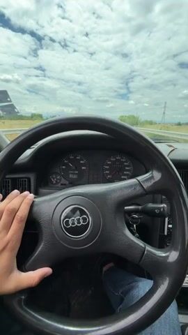 This is a first-person point-of-view video from inside a car. The driver's hand is visible on the steering wheel, which has an Audi logo in the center. The dashboard displays the speedometer and other gauges. The road ahead is visible through the windshield, with a cloudy sky and power lines in the distance. In the second half of the video, the camera pans to the passenger seat, where a young woman is sitting and holding a flyer or pamphlet. She turns to look at the camera with a neutral expression. The environment appears to be a highway in a rural or semi-rural area.