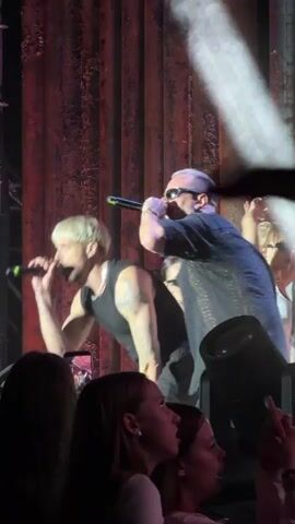 At 00:00, a performer with light-colored hair and sunglasses, wearing a dark sleeveless shirt and a black vest, sings into a microphone on a dimly lit stage. A large white spotlight shines on them from the right. In the foreground, the top of the heads of several audience members are visible, some holding up phones. At 00:01, the video shifts to an out-of-focus shot of a dark, possibly fuzzy surface. At 00:02, the video is still out of focus and dark. At 00:03, the performer is seen from the back, their arm raised with their finger pointing. Blue stage lights are visible. At 00:04, the performer, still wearing sunglasses and a black shirt, sings into a microphone and points with their left hand. A large inflatable white ball is in the upper left of the frame. Red stage lights are now visible, and the background appears to be a textured, reddish-brown curtain or wall. The audience is still visible in the foreground.
