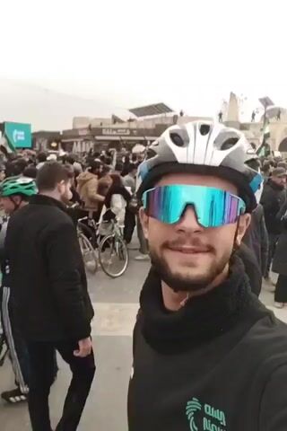 A man is taking a selfie in a crowd of people. The man is wearing a black sweater, a black scarf, a white and gray helmet, and reflective sunglasses. He is smiling. Behind him, there are other people, some of whom are wearing helmets and riding bicycles. There is a building in the background and flags and signs are visible. The weather appears overcast.