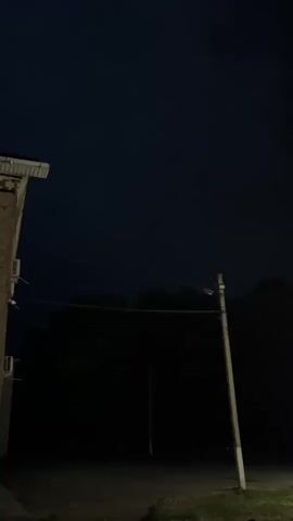 The video shows a dark, nighttime scene with a brick building on the left side. A utility pole with wires runs across the frame in front of a dark, silhouetted treeline. The sky is a deep blue-black. There are no people or overt actions visible.