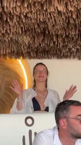 The video shows a woman with her hair pulled back in a ponytail, wearing a dark bikini top and a sheer white cover-up. She is looking up and to her right, smiling and raising her hands in a gesture of excitement. She is holding a pair of black headphones in her hands. Behind her, a thatched roof is visible, and to her left is a large, circular woven decoration. In the foreground, the shoulder and head of a man wearing a white shirt and glasses are visible. The woman is in what appears to be an outdoor or beach setting.