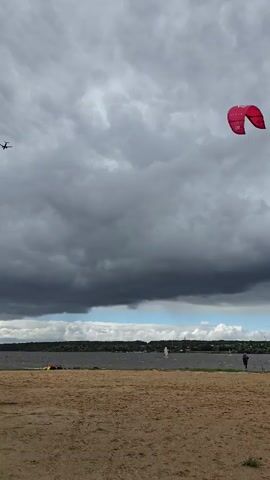 The video shows a sandy beach with a body of water in the background. The sky is overcast with dark clouds, and a small airplane is visible in the upper left corner. A red kite is flying in the upper right corner of the frame. There are a few people on the beach and in the water, some appearing to be windsurfing. A utility pole with a camera is in the right foreground.