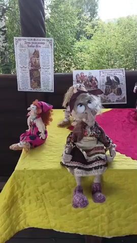 A group of dolls, some resembling witches with large noses and exaggerated features, are displayed on a table covered with a yellow and red cloth. The dolls are dressed in various outfits, some appearing to be handmade. Several informational posters with Russian text are visible in the background. The scene appears to be outdoors, with trees and foliage visible behind the table. The dolls are arranged in a somewhat scattered fashion, with some sitting and others standing. One doll is positioned near a small wooden house. The lighting suggests it is daytime.