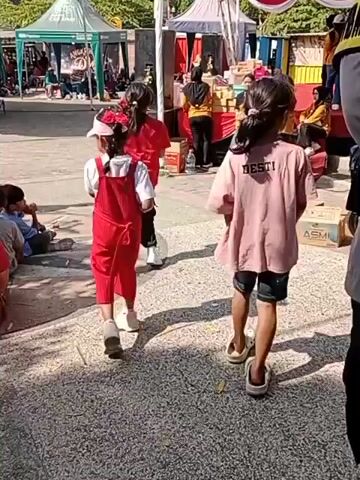 The video shows an outdoor event with several tents and people. In the foreground, four children are seated on the ground, facing away from the camera. Two children are standing and appear to be walking away from the camera, one wearing a red dress and the other a pink t-shirt. Behind them, a group of people are gathered around a stage-like area, with some individuals in yellow shirts. There are also several cardboard boxes visible on the ground. The environment appears to be sunny with shadows cast on the ground. The overall scene suggests a community event or festival.