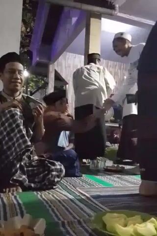 The video shows a group of men sitting on a patterned mat on the floor, likely indoors. One man in the foreground, wearing a checkered sarong, is holding a book. Another man, wearing a white shirt and pants with a white cap, is reaching out to shake hands with a man in a maroon shirt who is kneeling down. Several other men are visible sitting around them, some wearing traditional Islamic attire and caps. The environment appears to be a covered outdoor area or a veranda, with some lighting fixtures visible overhead. There are plates of food on the mat in the foreground.