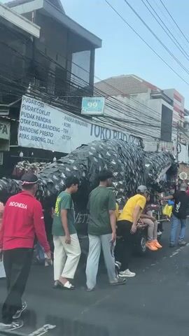 The video shows a street procession. A large, dark-colored dragon-like effigy, constructed from what appears to be overlapping dark material, is being pulled on a trailer with wheels. Several young men are walking alongside and behind the effigy, some pushing or guiding it. One individual in a yellow shirt is seated on the trailer. Behind the effigy, a group of people wearing red shirts and red hats are marching, many holding decorative circular objects with red and white elements. Some individuals in the crowd are observing the procession. The street is lined with buildings, some with shops displaying signs. Power lines are visible overhead. The pavement is wet in places. The overall atmosphere suggests a festive or celebratory event.