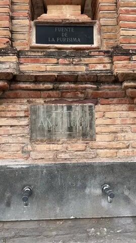 The video displays a close-up view of a brick structure that appears to be a historical fountain. At the top, a niche contains a statue of a woman with her hands clasped in prayer, adorned with white flowers. Below the niche, a dark plaque reads "FUENTE DE LA PURISIMA" in white lettering. Further down, a larger metallic plaque is inscribed with "AÑO 1881," indicating the year of construction. At the bottom of the structure, two silver spigots are visible, suggesting the fountain is functional. The brickwork is aged, with varying shades of orange and red, and the overall scene is well-lit, likely during daylight hours. The camera angle is mostly stationary, with slight upward movements, providing a detailed examination of the fountain's features.