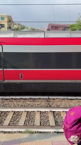 A train is parked at a station. The train is red and silver with "PREMIUM" written on it in white lettering. There are multiple doors on the side of the train, and one door is open. There are train tracks in the foreground, and a platform with a small building on it behind the tracks. The building is yellow and white with a closed gate. In the background, there are buildings on a hillside and trees. The sky is overcast.