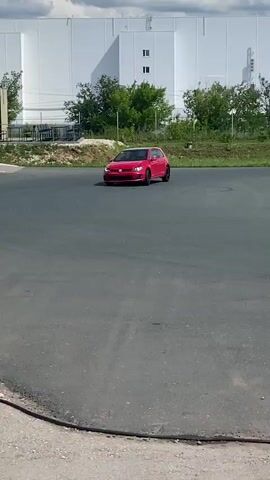 A red Volkswagen Golf GTI is seen driving around a tarmac circuit. The car is on the right side of the frame in the foreground and is moving right to left. Cones are placed around the circuit. In the background, there are trees, a building, and a cloudy sky. The car is filmed from a low angle.