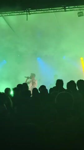 A man is singing into a microphone on a stage illuminated by green and blue lights. He is wearing a light-colored shirt. Behind him, another person is visible, possibly playing a guitar. A crowd of people, silhouetted in dark green, is gathered in front of the stage, watching the performance. The air is filled with a green haze.