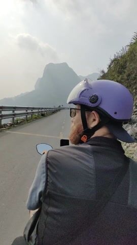 A man wearing a purple helmet, sunglasses, and a vest rides a scooter down a gray asphalt road. The camera angle is from behind the man, looking forward. A smartphone is mounted to the scooter. A gray metal guardrail runs parallel to the road on the left.  On the left of the road is water, and on the right is a rocky slope with greenery. In the distance, a large mountain is partially obscured by clouds. The sky is overcast.