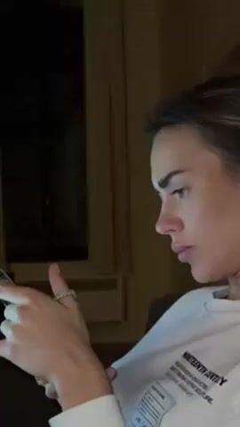 A woman is seen in profile, looking down and using a smartphone. She has light skin, dark eyebrows, and her hair is pulled back. She is wearing a white sweatshirt with black text on the front that reads "NINETEENTH CENTURY." On her left hand, she wears a ring with multiple small stones. The background is dimly lit, with a dark window and light-colored wall visible. The overall scene suggests a casual, indoor setting.