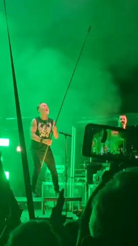 A man on stage holding a microphone attached to a long pole is seen singing and moving the pole. He is wearing a black t-shirt and black pants. Another man is standing behind him playing an instrument. The stage is lit with green lights, and there is smoke in the air. A crowd is visible in the foreground, some holding up their phones.