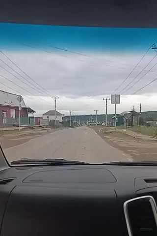 The video shows a view from the driver's seat of a vehicle traveling down a paved road through a rural village. Houses, some with fences, are visible on both sides of the road, along with utility poles and power lines. The sky is overcast with clouds. The vehicle's dashboard is visible in the foreground, including a display screen. The road ahead is mostly empty, with a few other vehicles sparsely scattered in the distance. The landscape appears to be somewhat hilly, with trees and vegetation visible in the background. The overall impression is of a slow drive through a quiet, residential area.