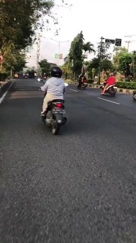 Four individuals are riding motorcycles in a line on a paved road. The rider in the front is wearing a dark jacket and helmet. The second rider is wearing a red jacket and helmet. The third rider is wearing a dark jacket and helmet. The fourth rider is wearing a camouflage jacket and helmet. The road is bordered by a sidewalk with trees and bushes. In the background, there is a traffic light and some buildings. The sky is visible, and it appears to be daytime.