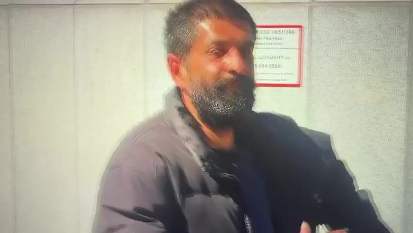 The video begins with a close-up of a man with a beard and dark jacket standing in front of a wall with a sign. The camera then cuts to a medium shot of a person wearing a mesh tank top and a headband, smiling and giving a thumbs-up. They are standing next to a bus with a yellow sticker that reads "Beware of passing this vehicle on the inside." The video then transitions to a wide shot of a street scene with people walking on a sidewalk in front of modern buildings. A black pillar stands prominently in the foreground.