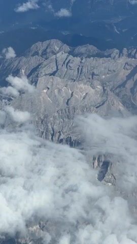This is a high-angle aerial shot of rugged, jagged mountain peaks partially obscured by soft, white clouds. The mountains appear to be rocky with steep slopes and deep crevices. Sunlight casts shadows across the landscape, highlighting the textures and contours of the rock formations. In the distance, the terrain becomes less distinct, blending into darker, forested slopes under a hazy blue sky. The overall impression is one of a vast, natural mountain range viewed from a great height. There are no people or man-made objects visible.