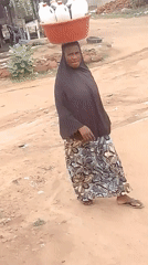 Woman carries red basin with containers while walking dirt road