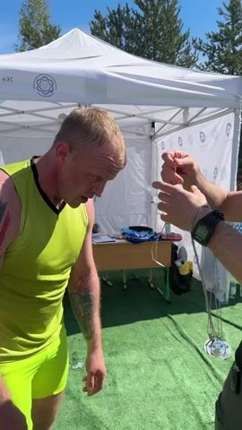 The video shows a man being awarded a medal. The man is wearing a yellow tank top and shorts. He is standing in front of a white tent. The person awarding the medal is standing in front of him. He is wearing a black t-shirt and sunglasses. The man accepting the medal has tattoos on both arms. After receiving the medal the two men shake hands.