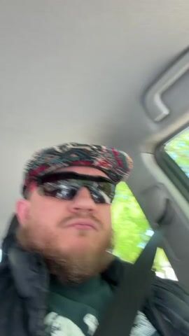 A man with a beard, wearing a patterned cap and sunglasses, is driving a car. He has a serious expression on his face. He is wearing a black vest over a green t-shirt and has his seatbelt fastened. The interior of the car is visible, including the headliner, window, and side panel. Trees are visible outside the car window. Music is playing.