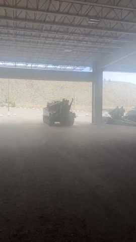 The camera pans from left to right, under a roofed structure with steel support beams.  Outside the roof, there is bright sunlight and a hillside.  Inside the structure, various military vehicles are parked on dirt.  The camera focuses on a camouflage military truck.